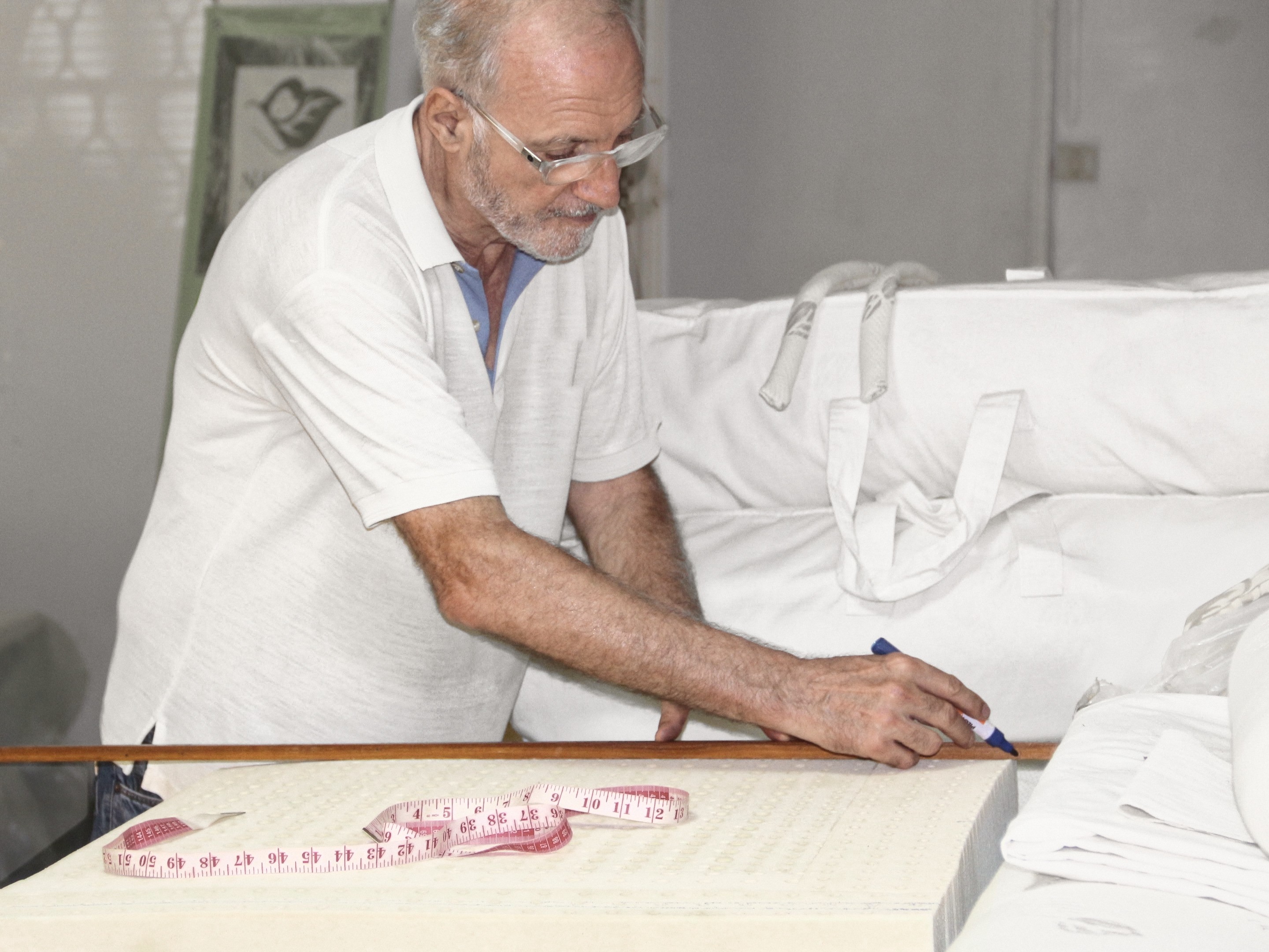 Dominique Bienfait in a white polo and glasses, bent over a block of latex on his work table, drawing a line with a marker along a wooden rule, a tape measure lying on the block and white cotton covers stacked behind him.