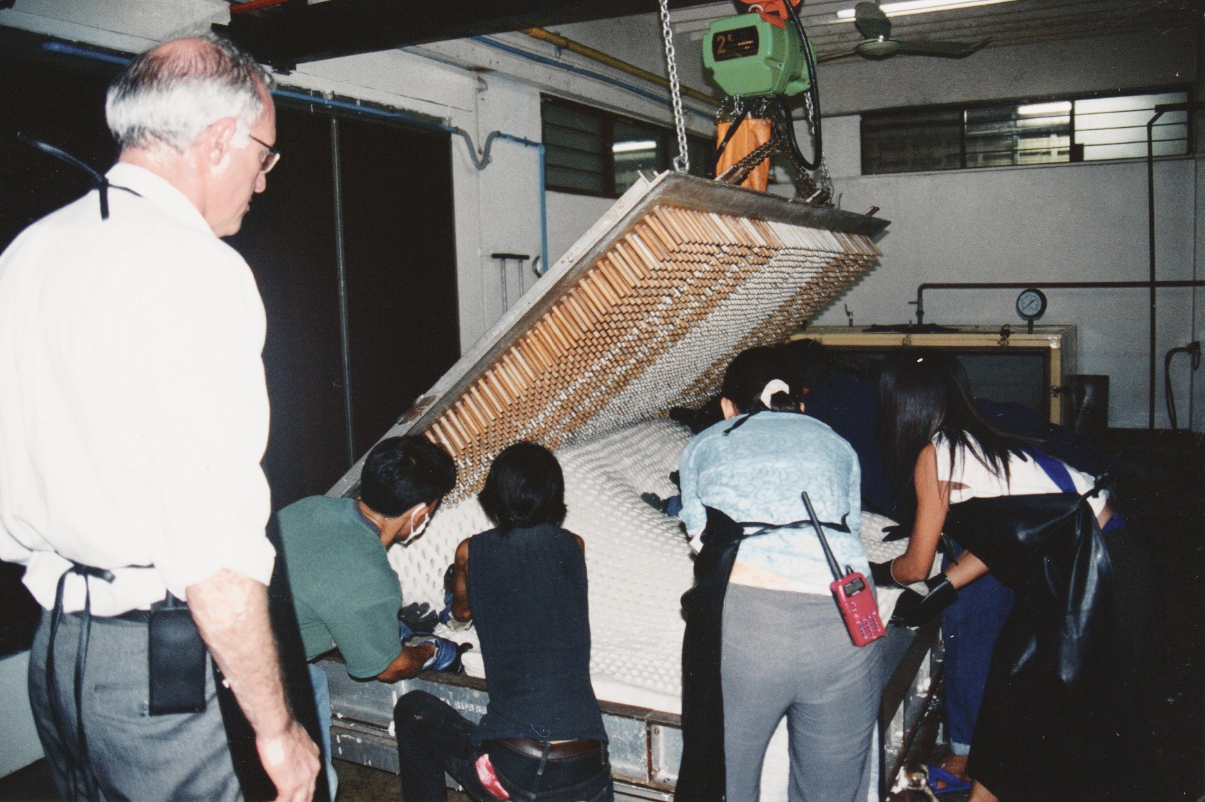 An old print in a latex factory: a mould of long pins raised on a hoist over a white latex core, four workers leaning over the core and Dominique Bienfait watching from the left.