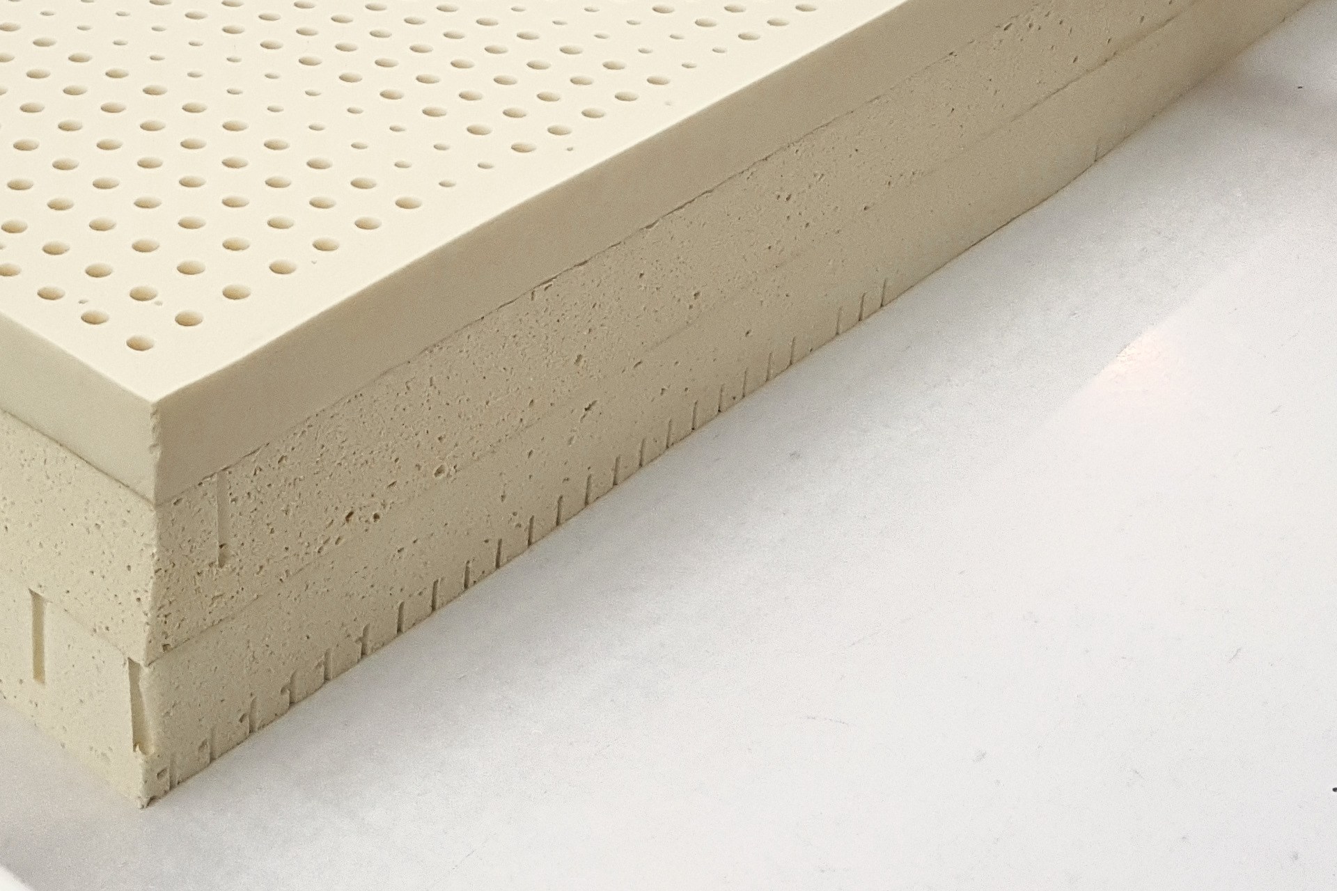 The near corner of a stack of three slabs of latex on a white table: the smooth perforated top, the open cells of the cut sides, a row of slits along the lower edge.