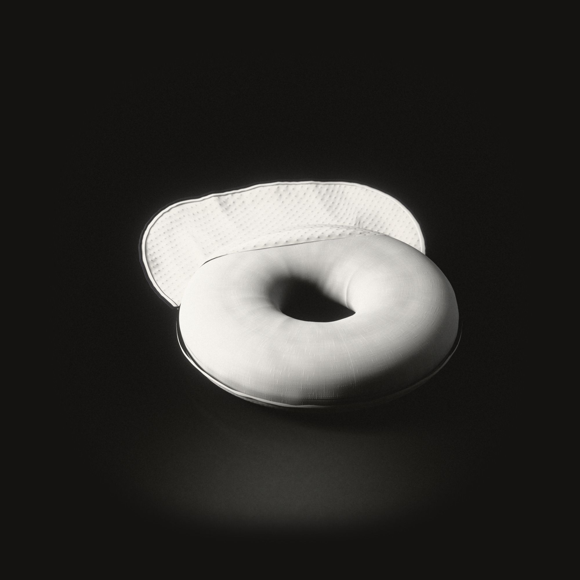 Un coussin blanc en forme d’anneau sur un sol noir, sa housse tricotée à pois ouverte et rabattue derrière lui.