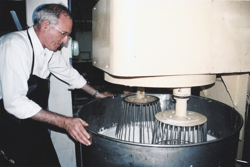 An old print: a man with white hair, glasses and a black apron, his hand on the bowl of a foaming machine whose two wire whisks turn in white latex.