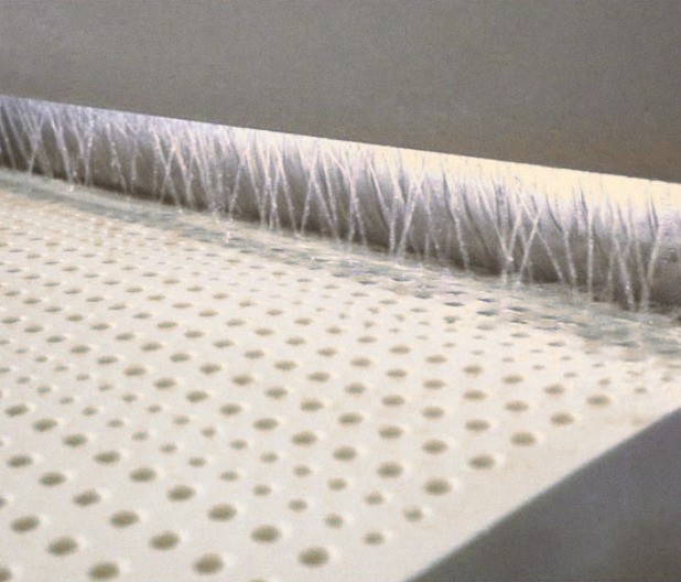 A block of perforated latex passing under a curtain of water jets, the water running over its rows of pin holes.
